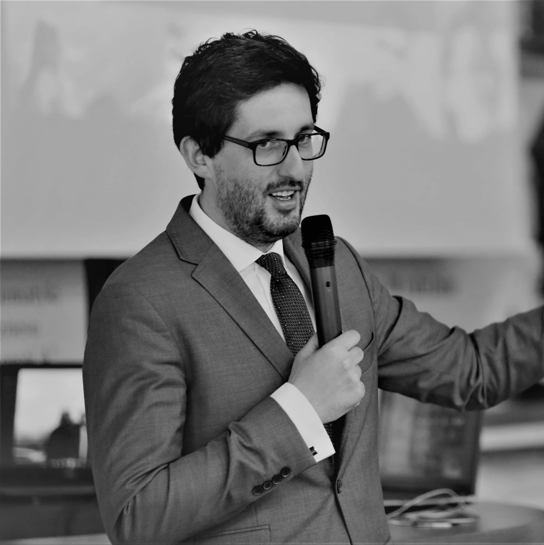 Mężczyzna w garniturze trzyma mikrofon i gestykuluje podczas prezentacji.