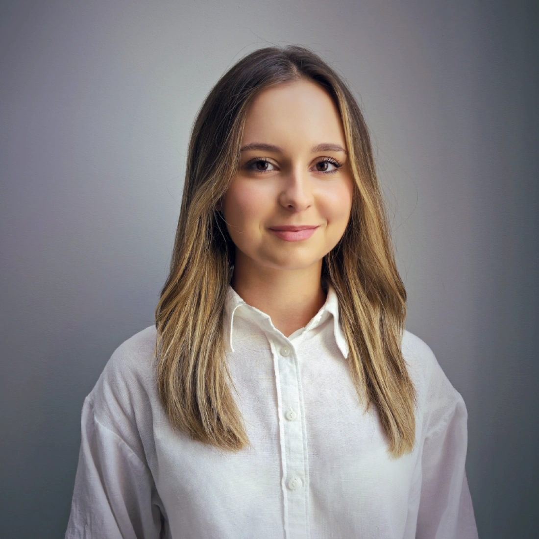 Portret młodej osoby w białej koszuli, z długimi, prostymi włosami. Tło jest jednolite i jasne.