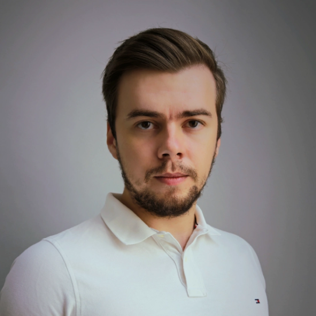 Mężczyzna z krótkimi włosami i brodą, ubrany w biały podkoszulek, patrzy w obiektyw.