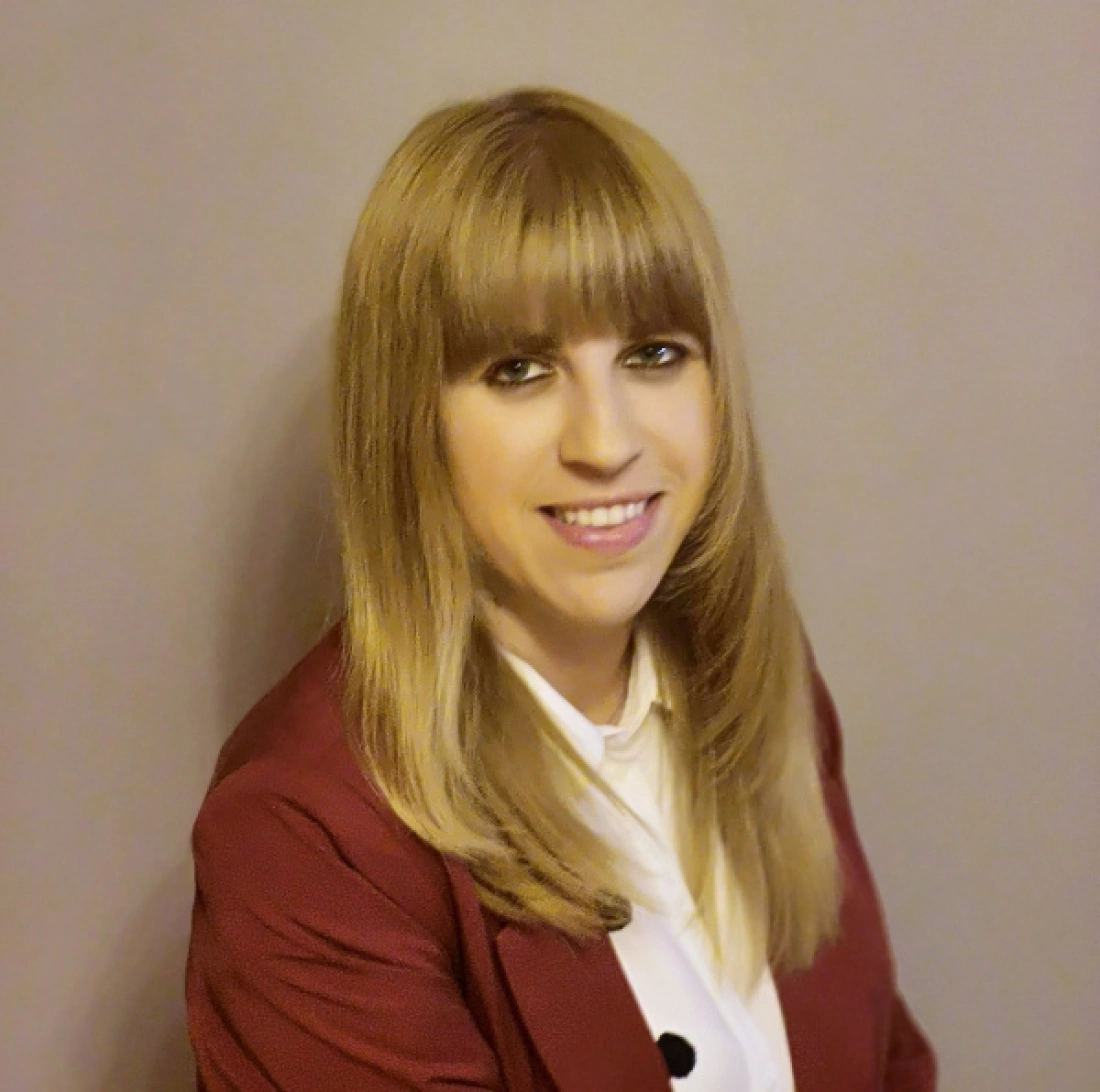 Osoba z długimi, blond włosami, ubrana w czerwoną marynarkę i białą koszulę, uśmiecha się na tle szarej ściany.