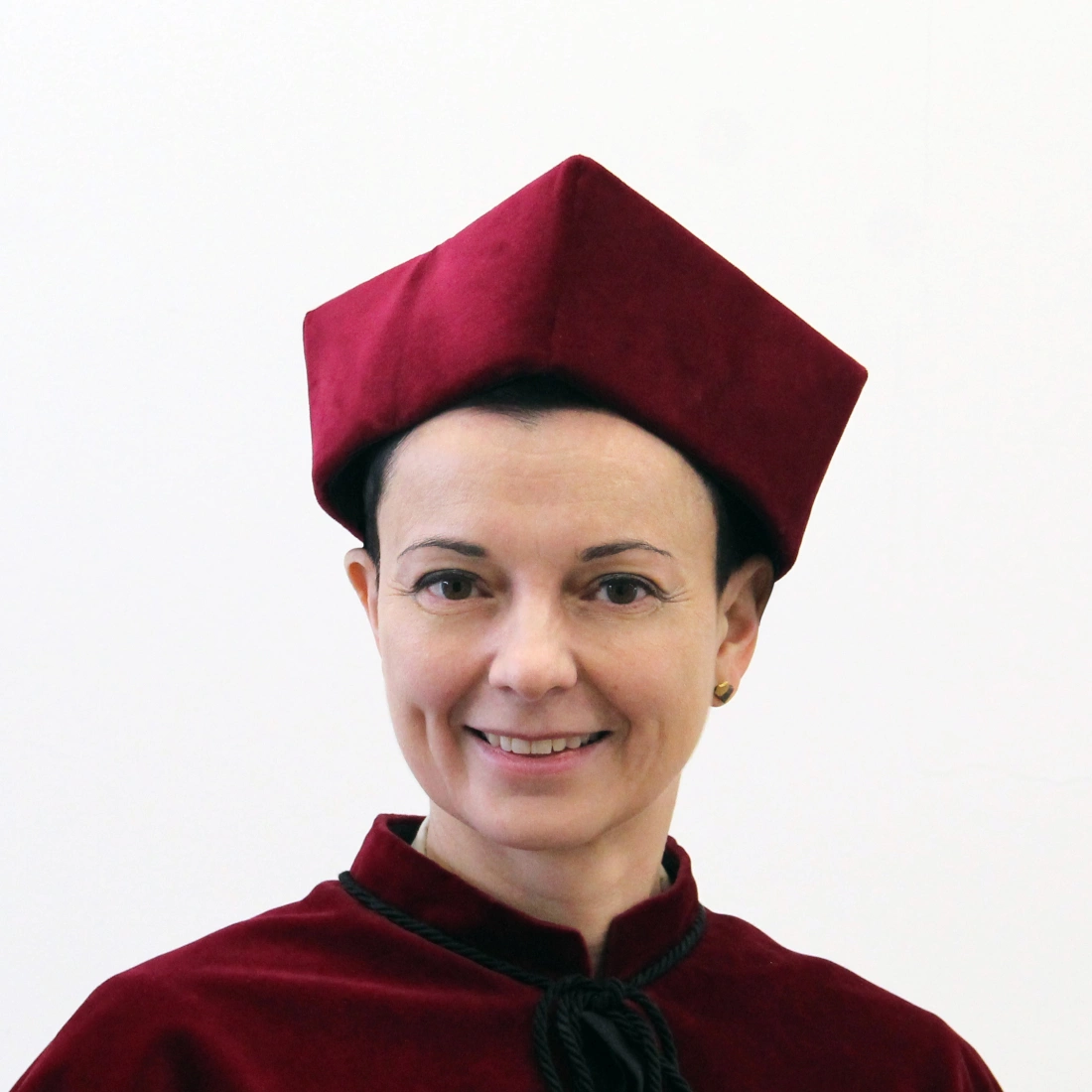 Kobieta w burgundowym stroju akademickim z uśmiechem na tle jasnej ściany.