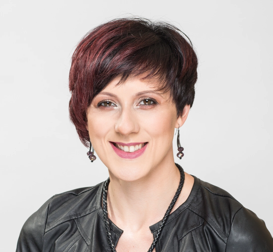 Portret uśmiechniętej kobiety z krótkimi włosami w odcieniach czerwieni, w skórzanej kurtce i biżuterii.