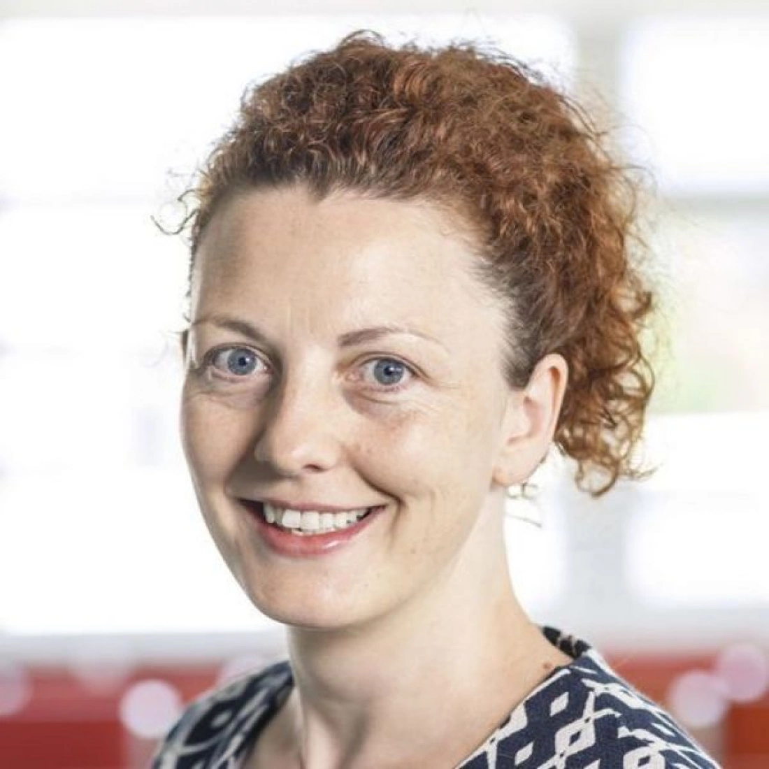 Uśmiechnięta kobieta z kręconymi, rudymi włosami, w stonowanej bluzce, stojąca w jasnym otoczeniu.