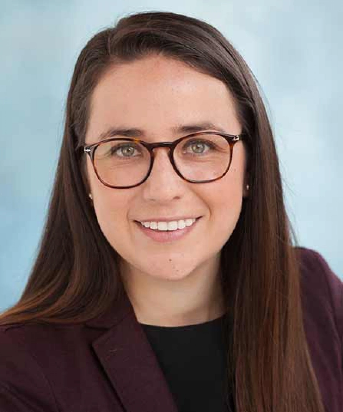 Portret uśmiechniętej kobiety w okularach, z długimi, prostymi włosami, w ciemnym garniturze na tle w odcieniach niebieskiego.