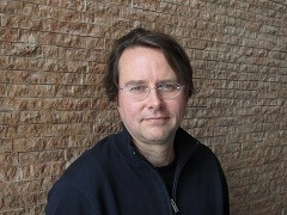 Mężczyzna w okularach, stojący przed ceglanym tłem.