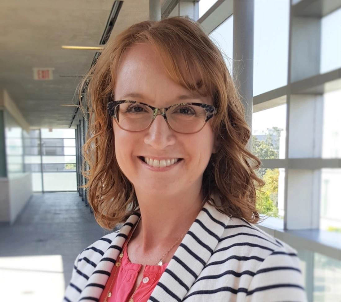 Uśmiechnięta kobieta z kręconymi włosami, w okularach i wstripowanej marynarce, stoi w jasnym korytarzu.