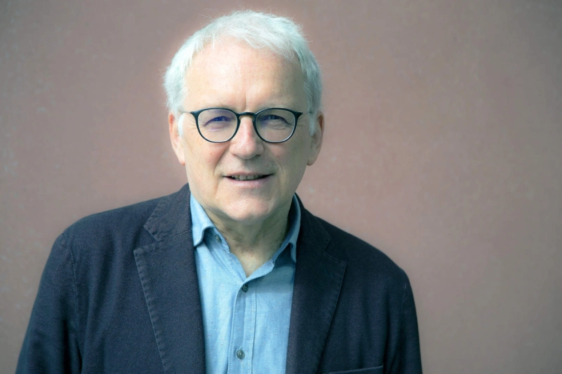 Mężczyzna z siwymi włosami i okularami, uśmiechający się na tle ściany.