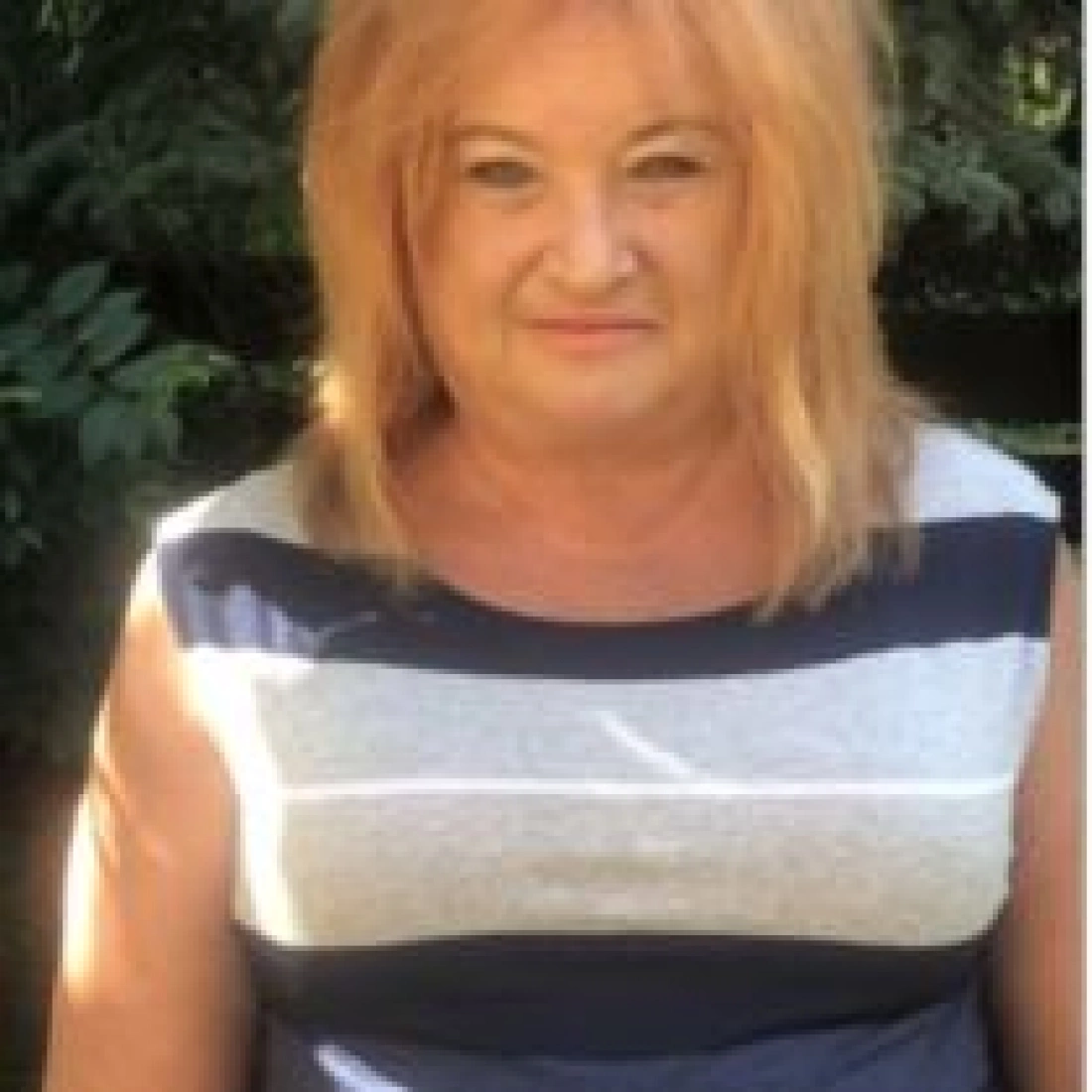 Kobieta z długimi, blond włosami, ubrana w bluzkę w poziome paski, stojąca na tle zieleni.