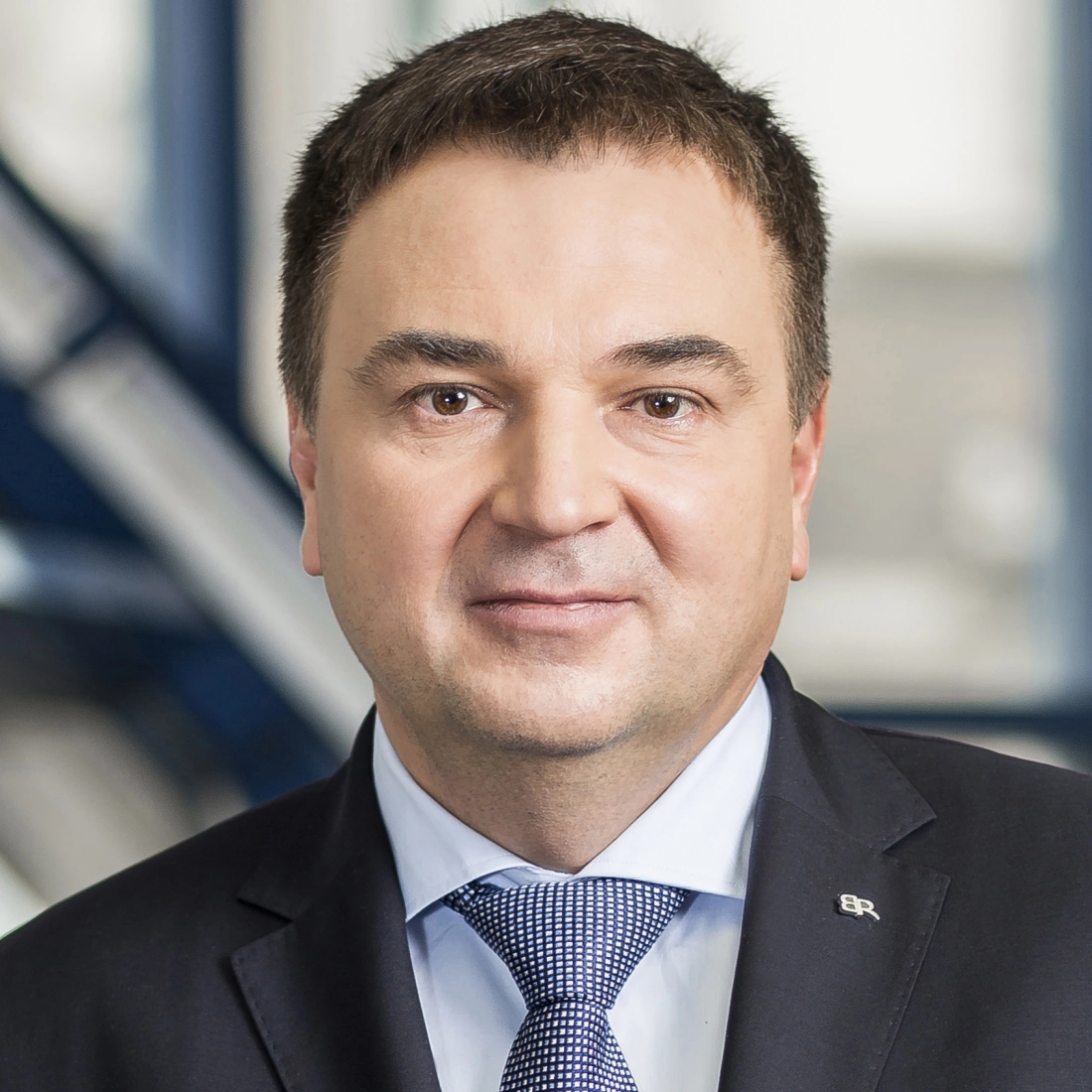 Portret mężczyzny w garniturze, z delikatnym uśmiechem, na tle nowoczesnego wnętrza.