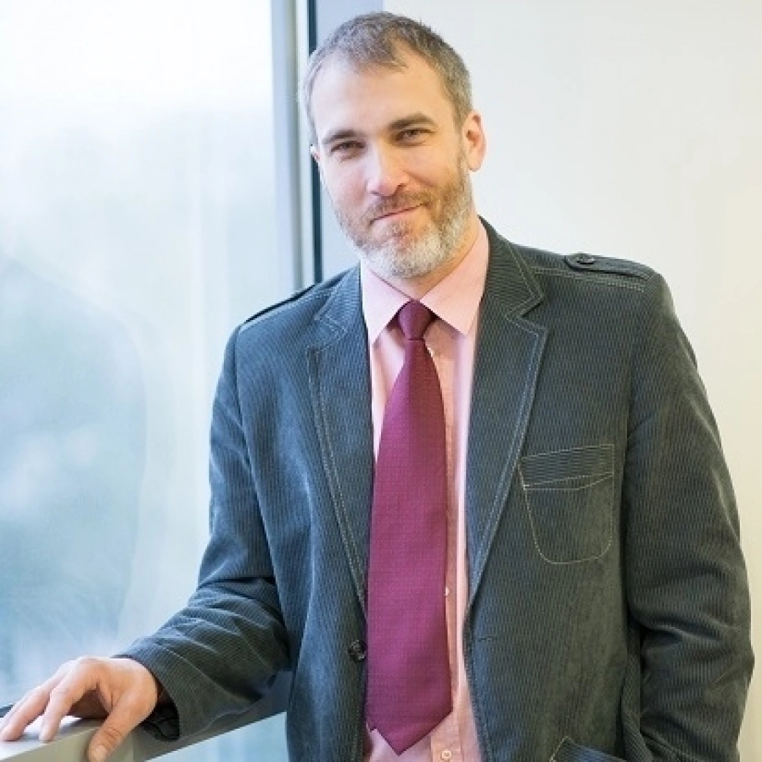 Mężczyzna w eleganckim garniturze stoi przy oknie, lekko się uśmiechając.