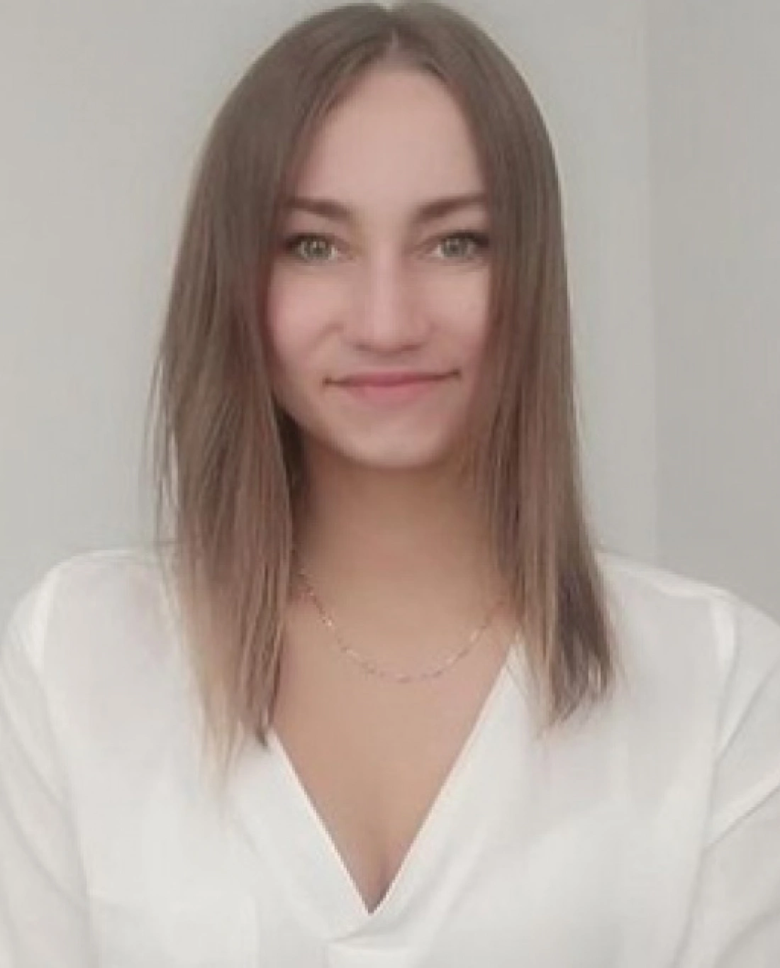 Młoda kobieta uśmiecha się, nosząc białą koszulę, na tle jasnej ściany.