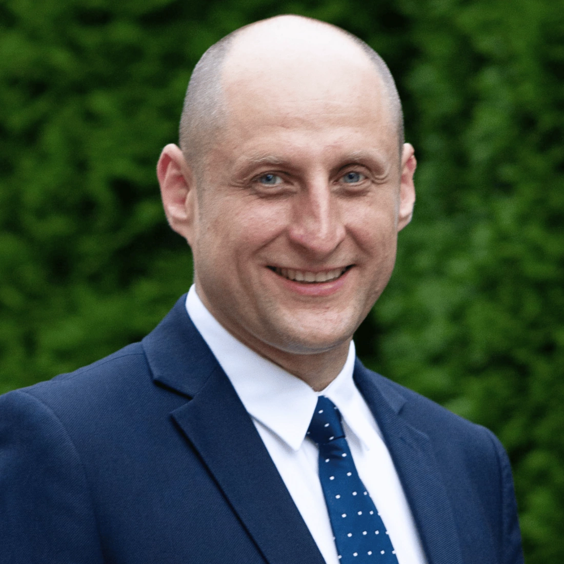 Mężczyzna w garniturze z krótkimi włosami i niebieskimi oczami, uśmiechający się do kamery.