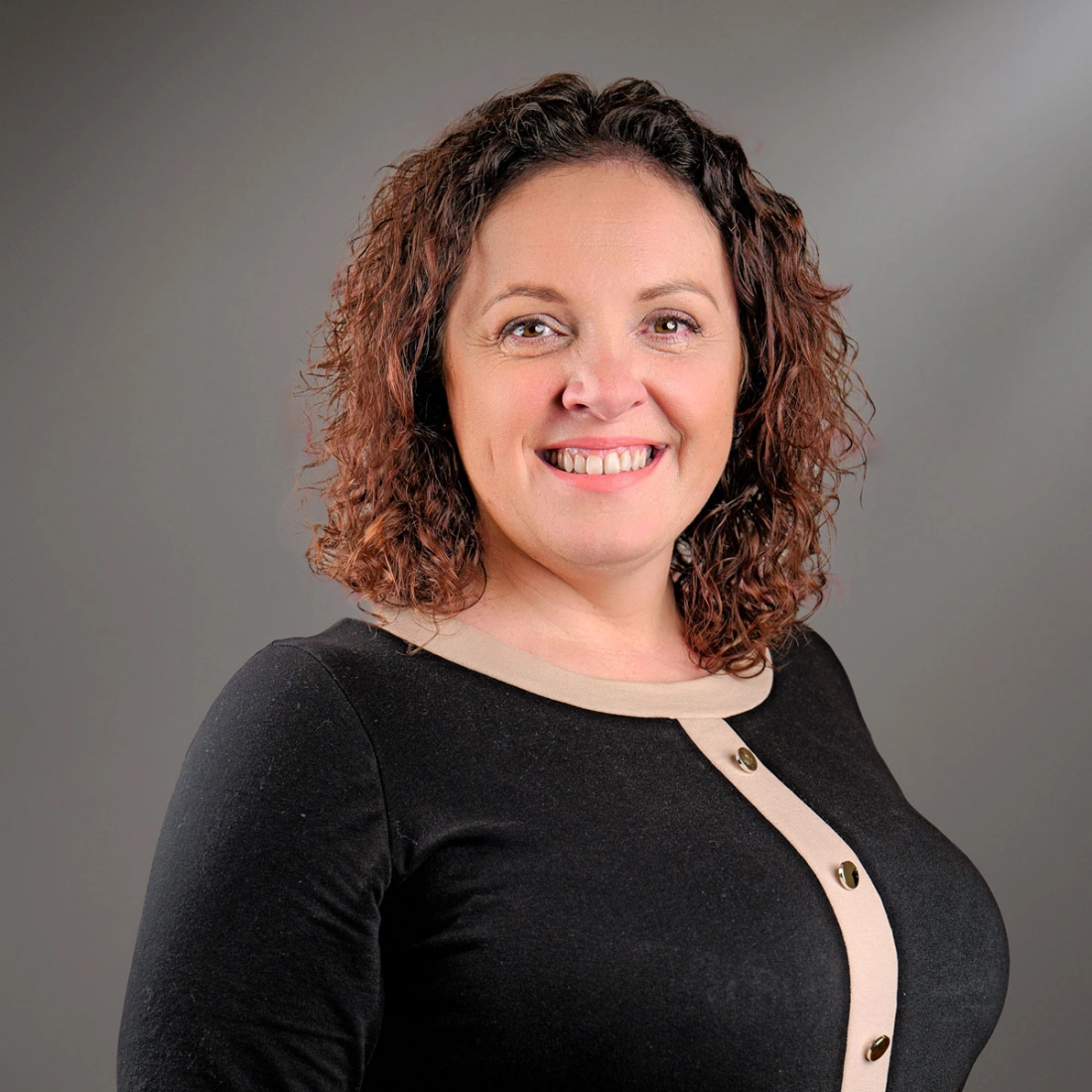 Kobieta z kręconymi czerwonymi włosami, uśmiechnięta, w bluzce w jasnym kolorze, na tle zieleni.