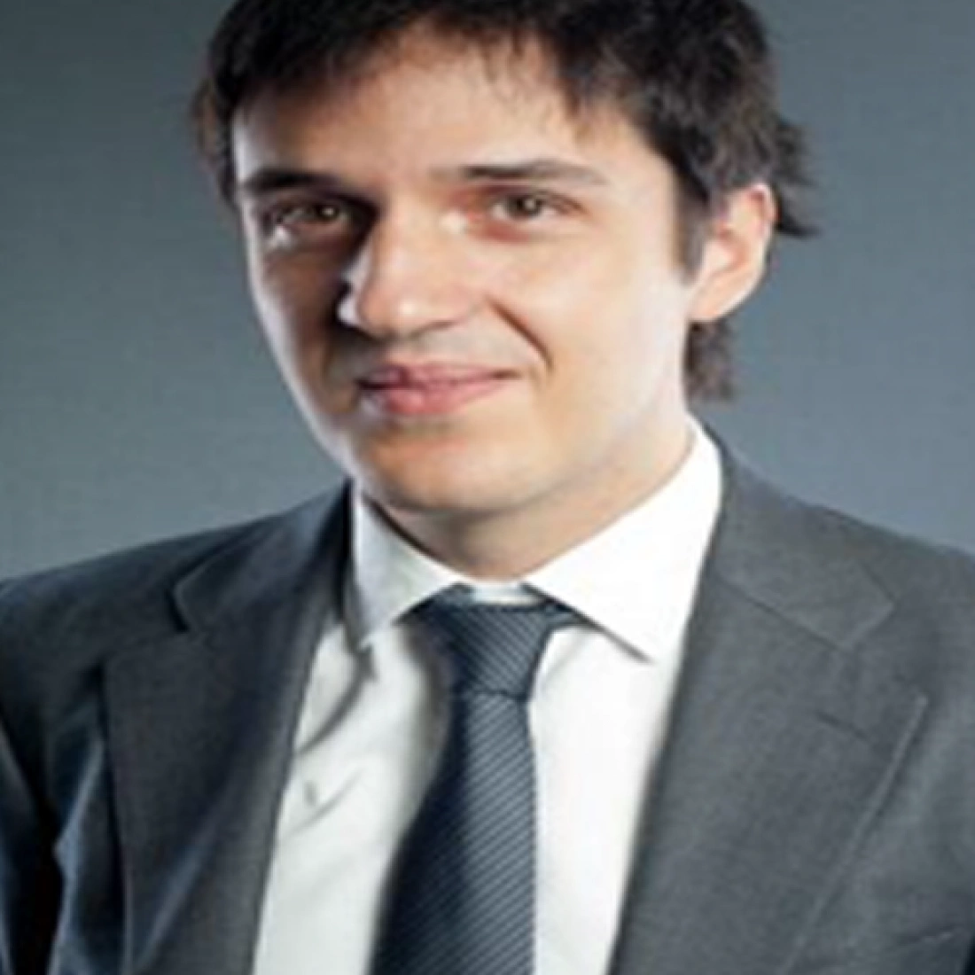 Mężczyzna w szarym garniturze z uśmiechem, na tle jasnej ściany.