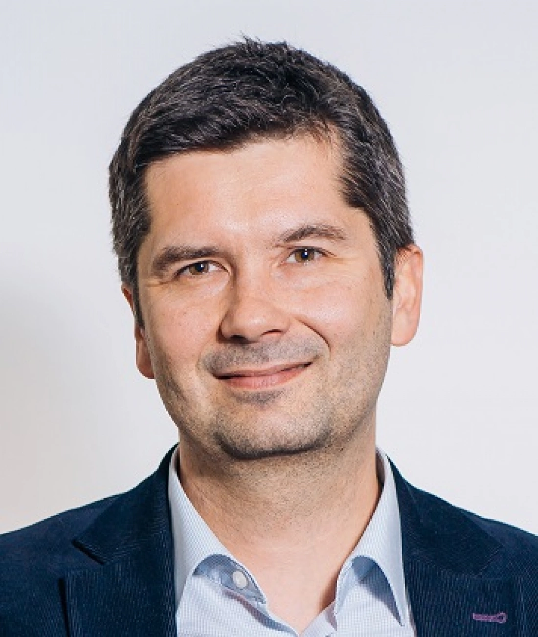 Mężczyzna w ciemnym garniturze uśmiecha się, z jasnym tłem za sobą.