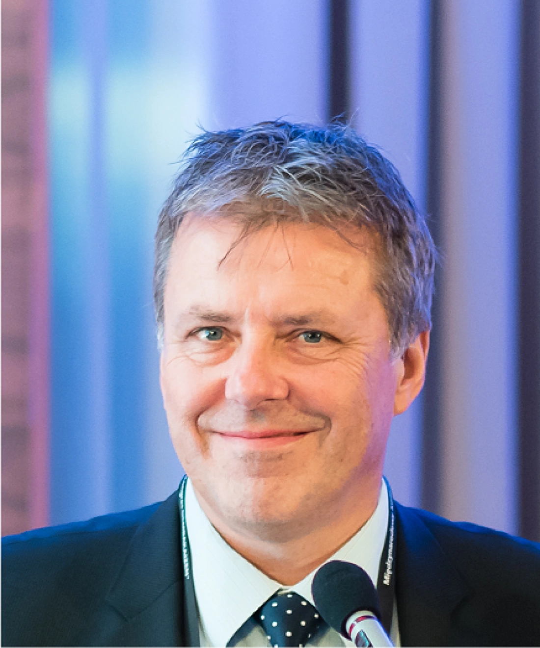 Mężczyzna w garniturze z uśmiechem, stojący przy mikrofonie.