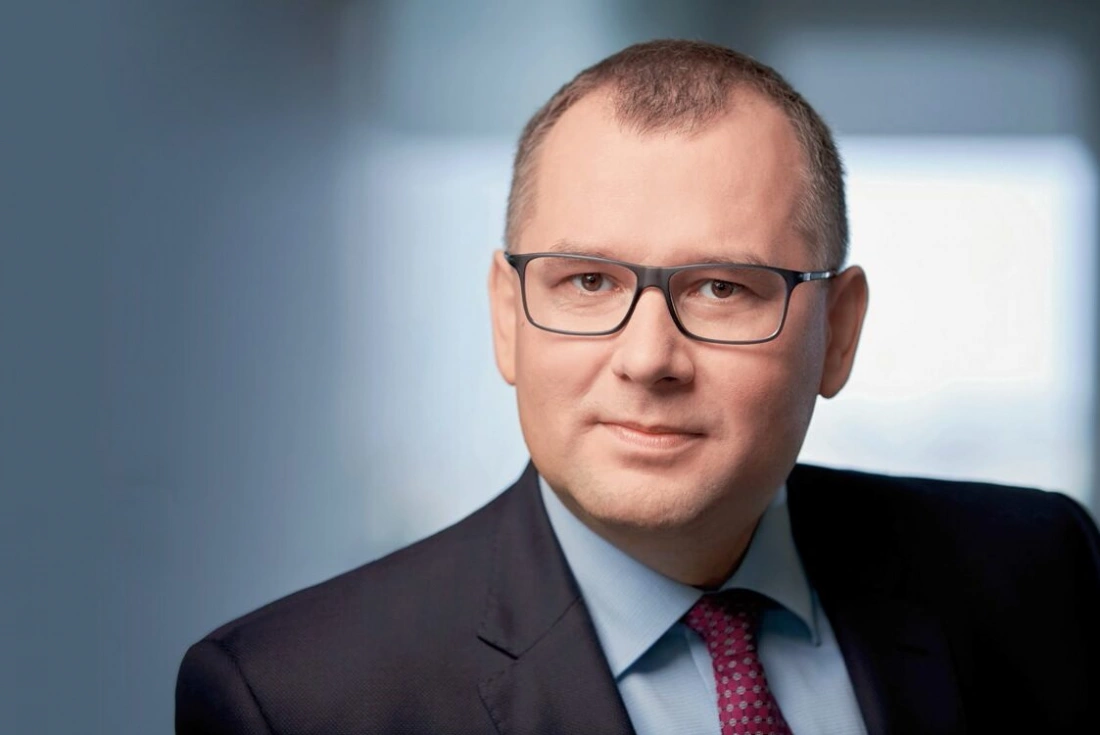 Mężczyzna w garniturze z okularami, z poważnym wyrazem twarzy, na tle rozmytej przestrzeni.