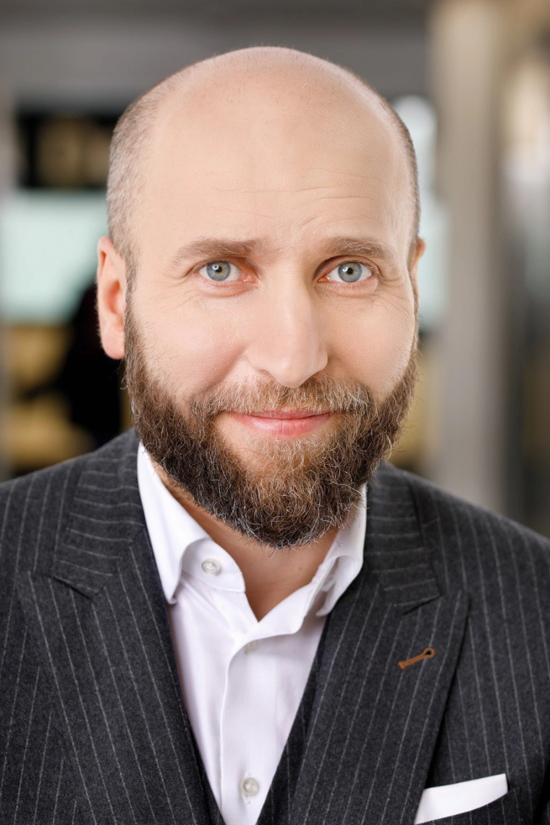 Mężczyzna z brodą, w eleganckim garniturze, uśmiecha się do aparatu.