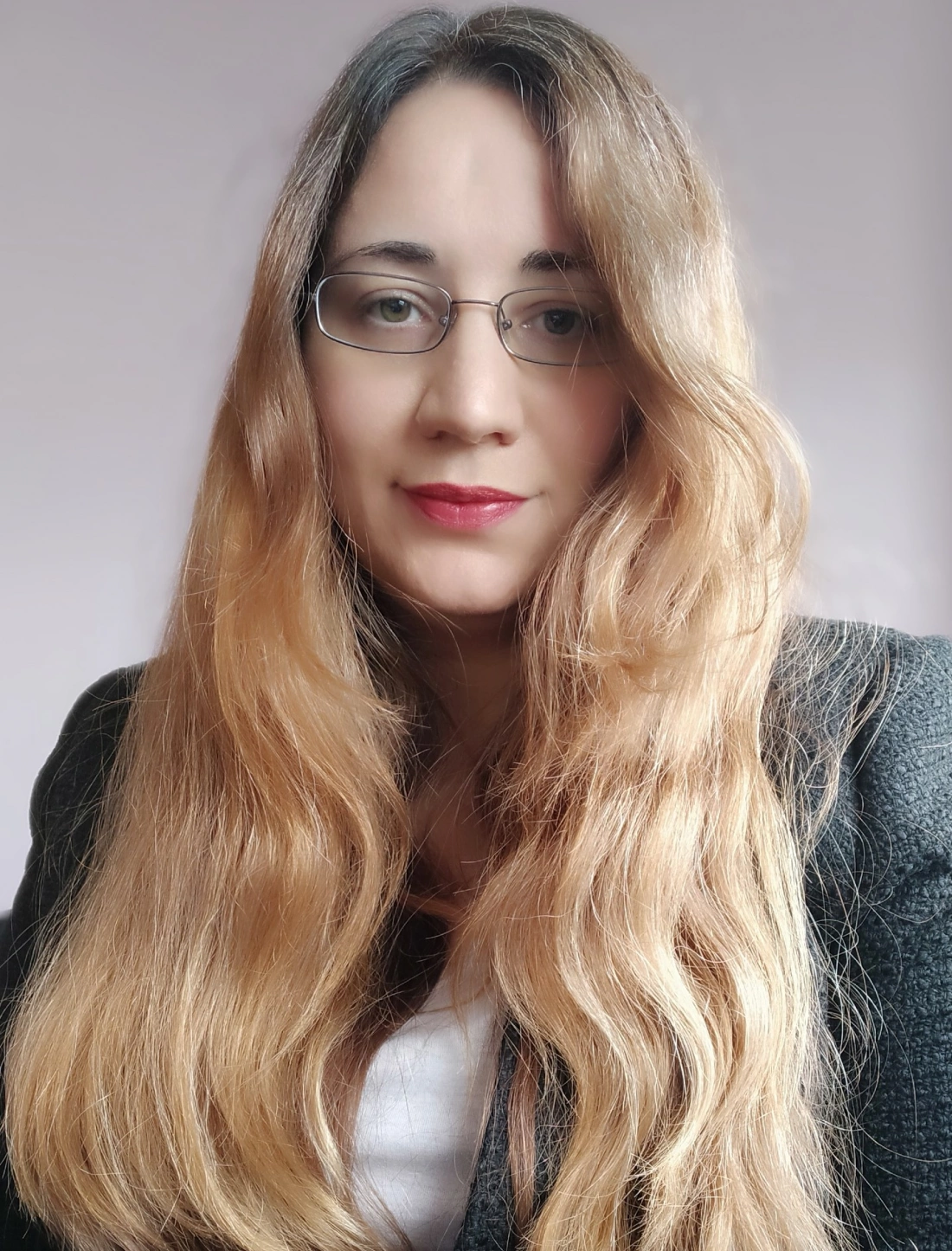 Portret kobiety z długimi, falującymi włosami, w okularach, z delikatnym makijażem, ubranej w ciemny żakiet. Tło w jasnym kolorze.