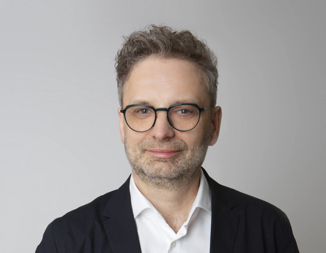 Mężczyzna w okularach, ubrany w czarny żakiet i białą koszulę, uśmiechający się na szarym tle.