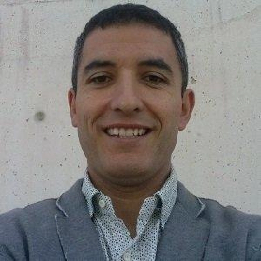 Mężczyzna uśmiechający się do aparatu, w jasnym garniturze i koszuli w wzorki, na tle gładkiej ściany.