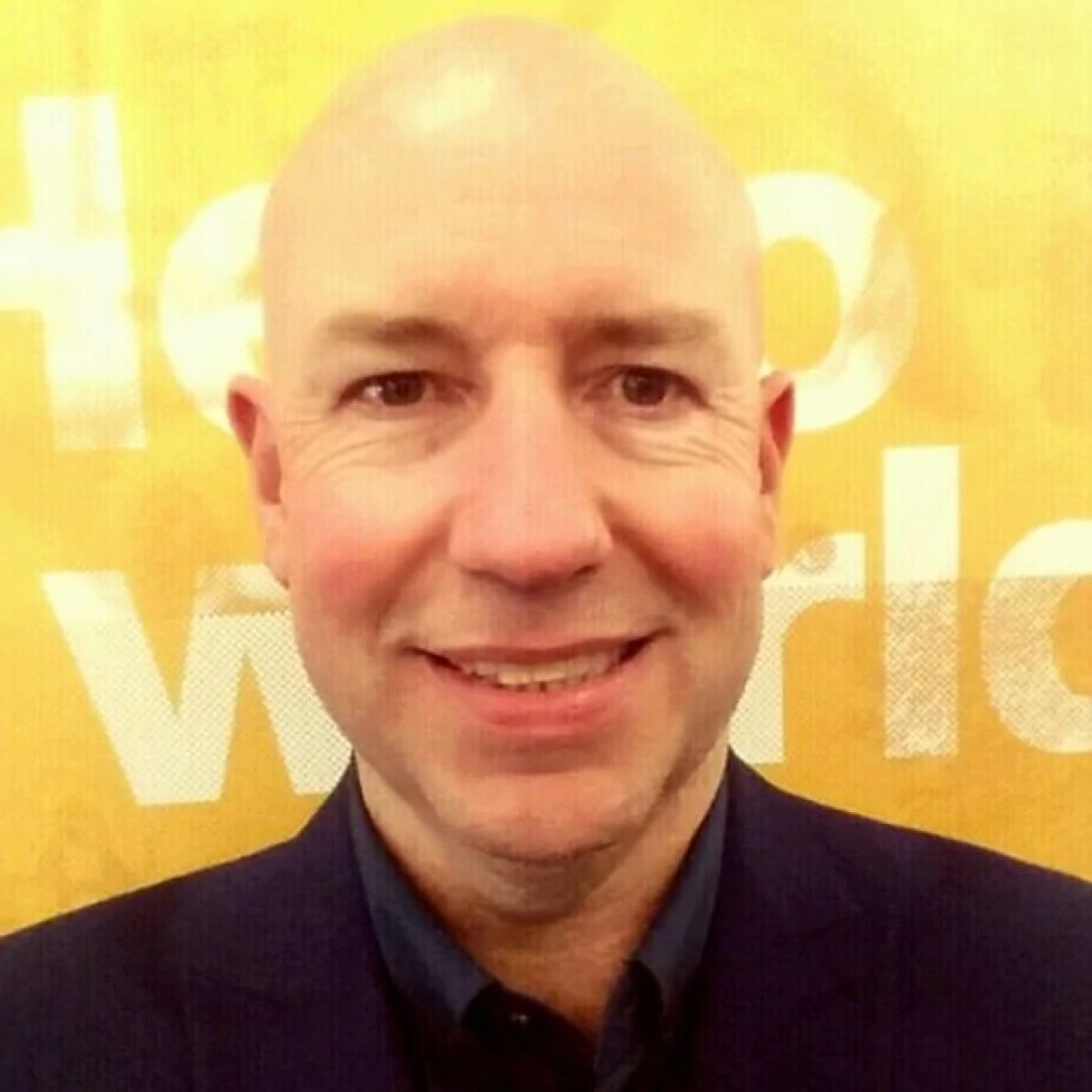 Mężczyzna z łysiną w garniturze, uśmiechający się przed żółtą ścianą z napisem.