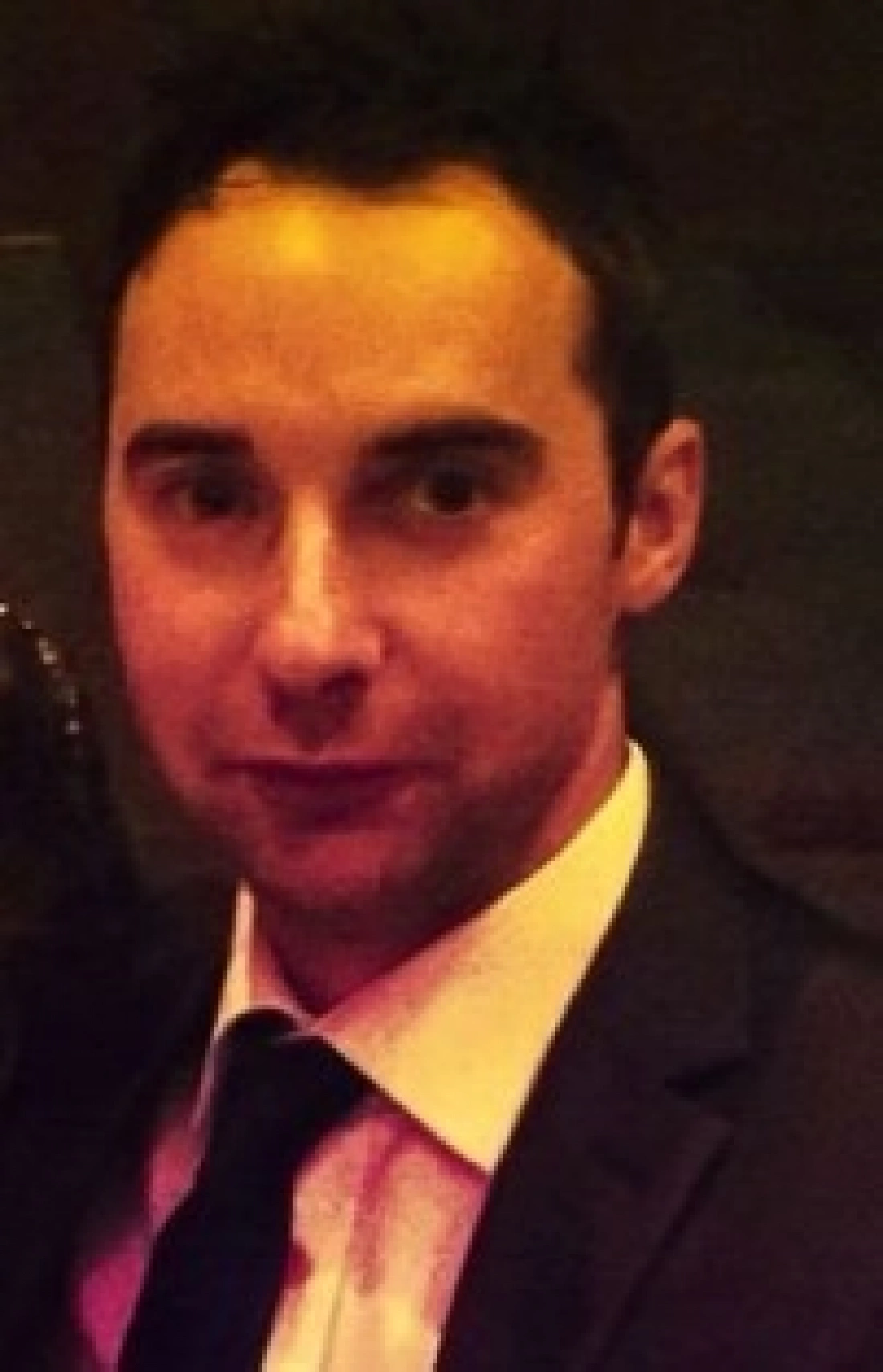 Mężczyzna w garniturze, z krótkimi, ciemnymi włosami, patrzy w obiektyw.