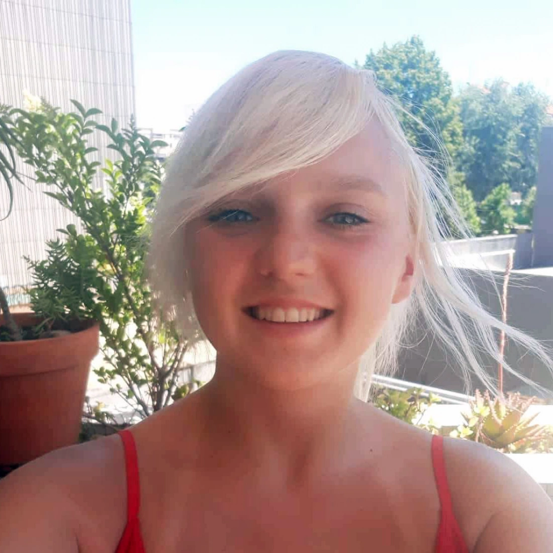 Młoda osoba z blond włosami uśmiecha się na tle roślin i nieba.