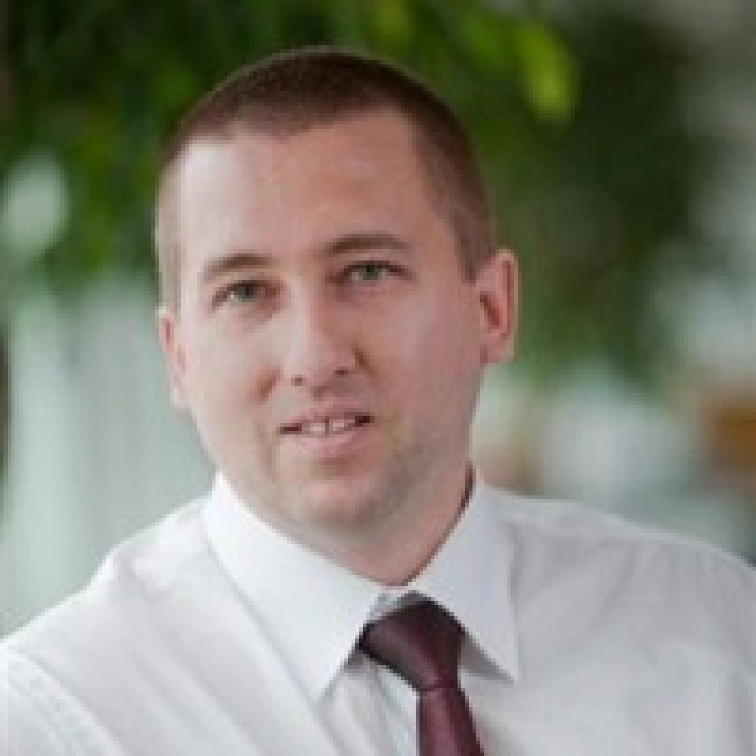 Mężczyzna w białej koszuli z krawatem, uśmiechający się na tle roślinności.