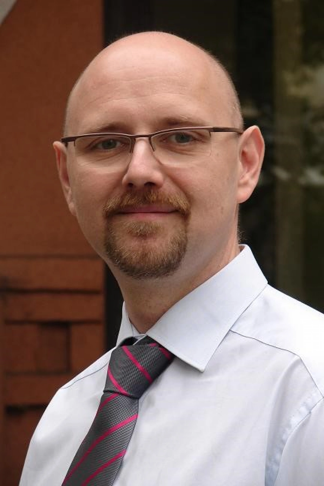 Mężczyzna w okularach, z brodą, ubrany w białą koszulę i kolorowy krawat, uśmiecha się na tle budynku.