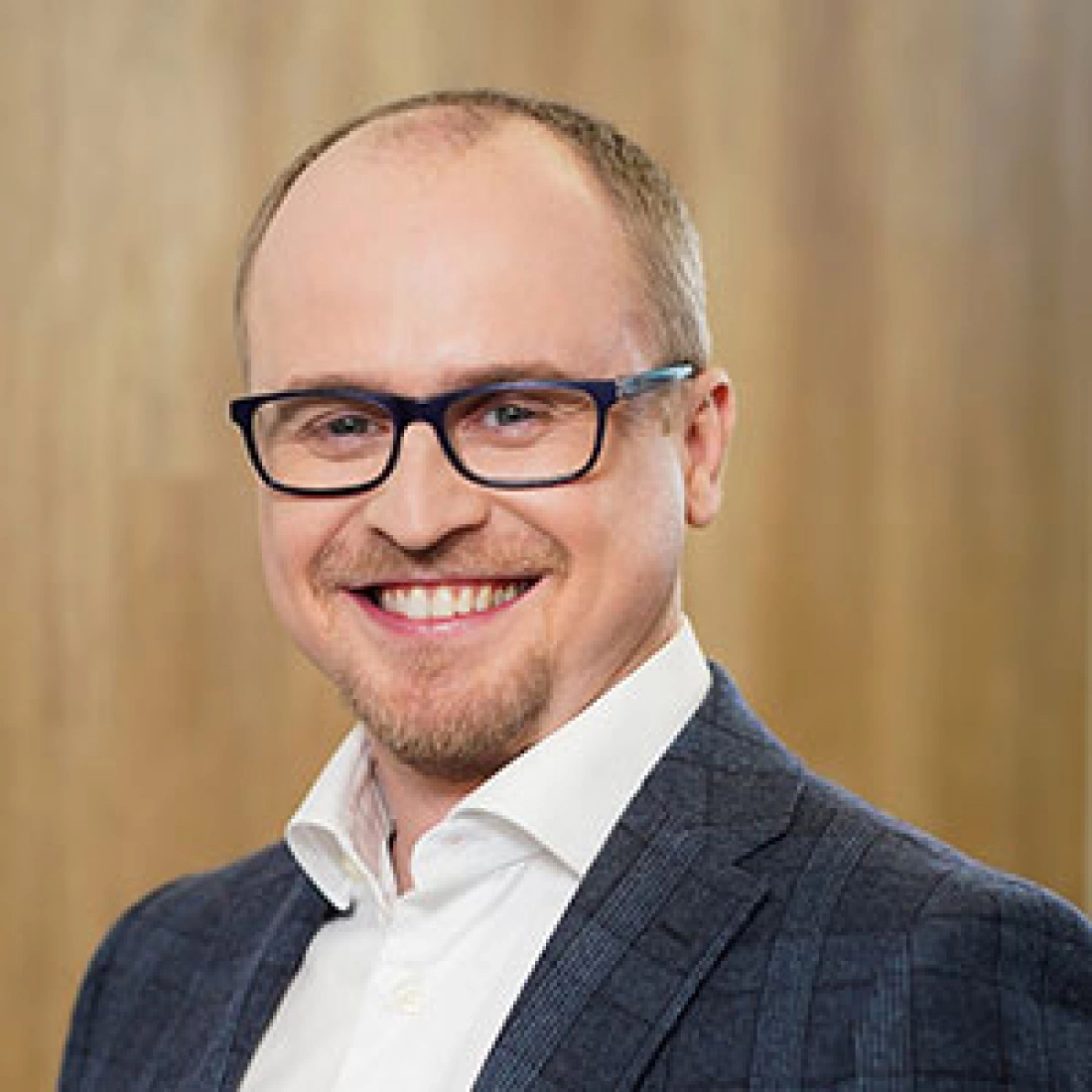 Mężczyzna w okularach, uśmiechający się w formalnym stroju na tle drewnianej ściany.