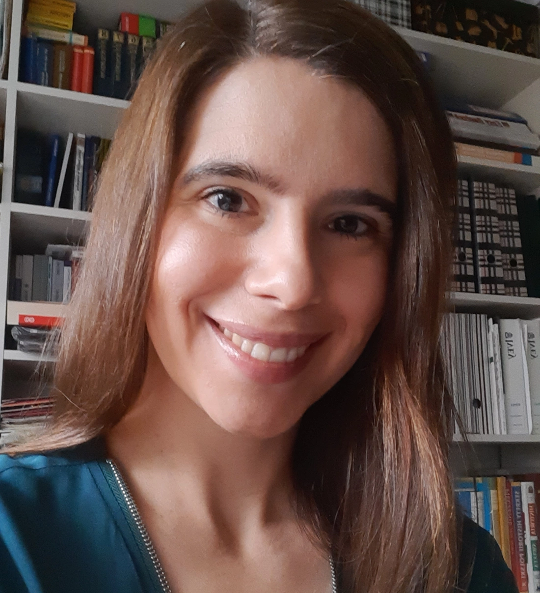 Młoda kobieta uśmiecha się do aparatu, w tle widać półkę z książkami i dokumentami.