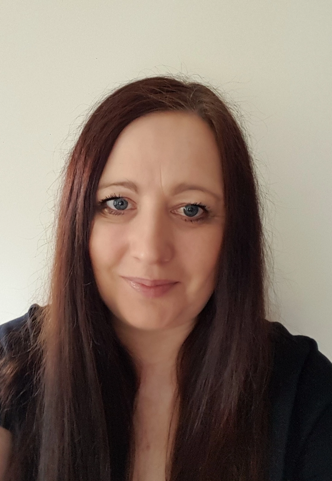 Kobieta z długimi, prostymi włosami, uśmiecha się do aparatu. W tle jasna ściana.