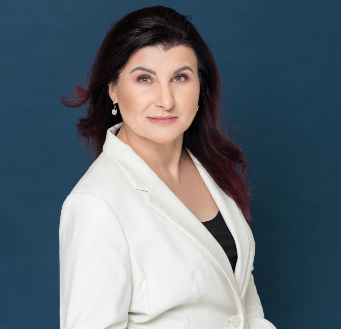 Kobieta w białym garniturze z długimi, ciemnymi włosami, pozująca na jasnoniebieskim tle.