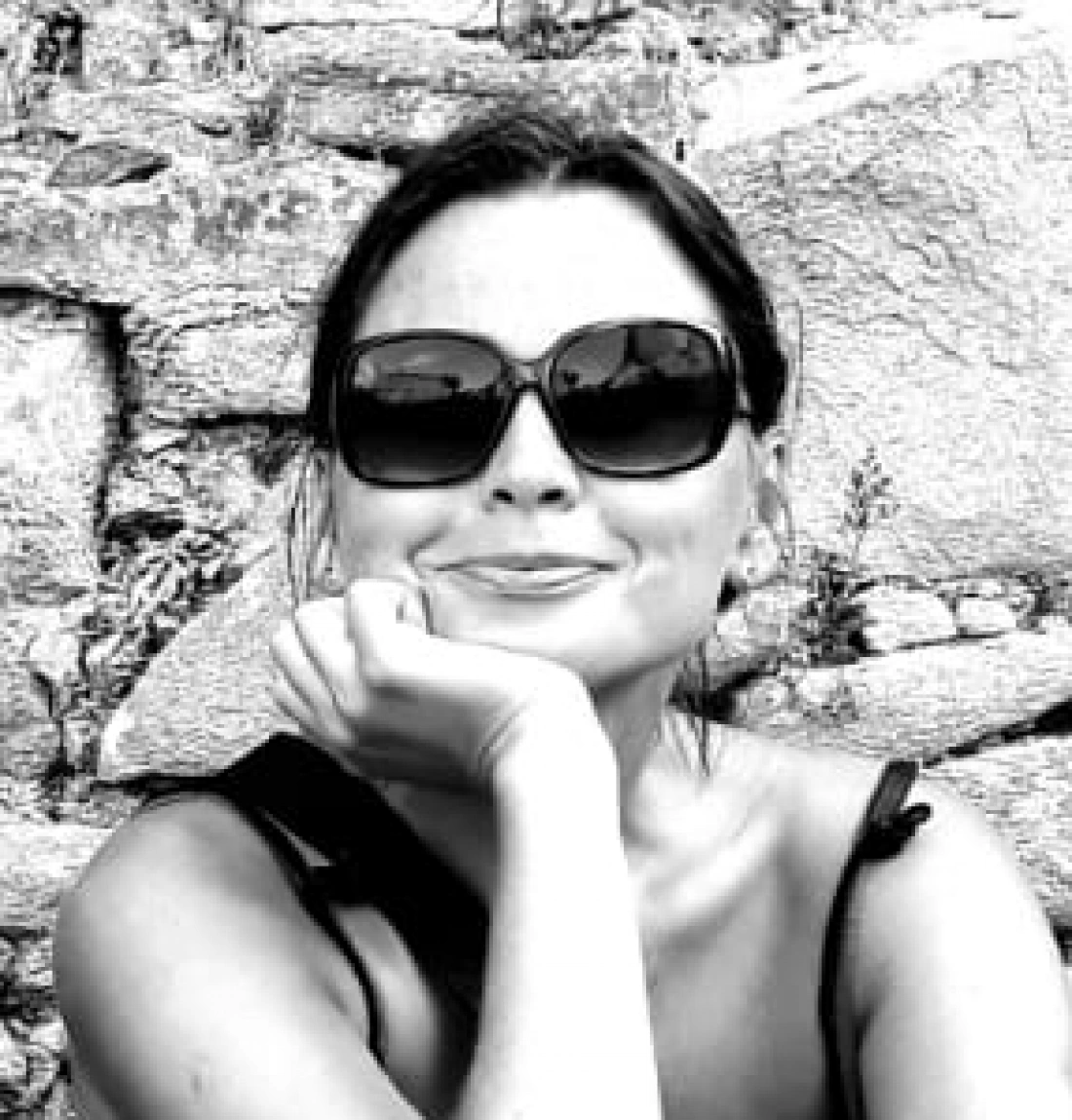 Kobieta w ciemnych okularach przeciwsłonecznych uśmiecha się, opierając głowę na dłoni, w tle kamienna ściana.