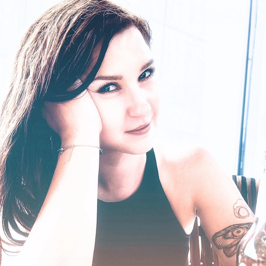 Młoda kobieta z tatuażem, uśmiechająca się przy dłoni na twarzy.