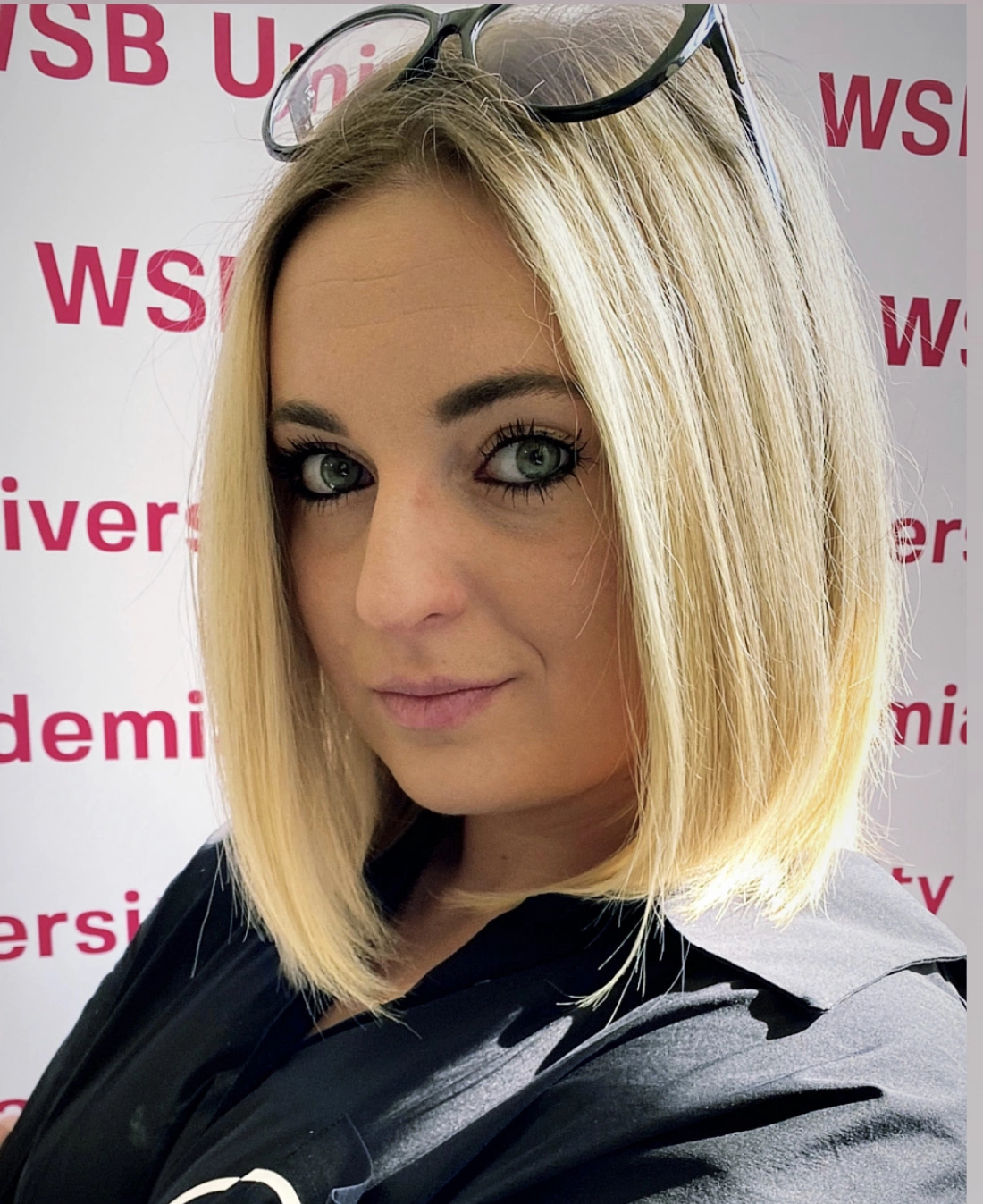 Portret kobiety z blond włosami, w czarnej bluzce, z okularami na głowie, stojącej przed tłem z napisem „WSB University”.