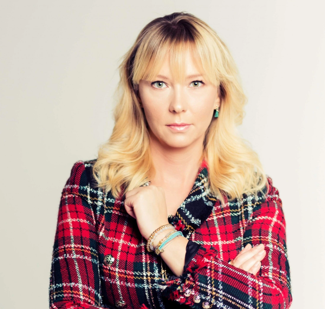 Młoda kobieta w czerwonym, w kratkę żakiecie, z poważnym wyrazem twarzy, trzyma ręce skrzyżowane na piersi.