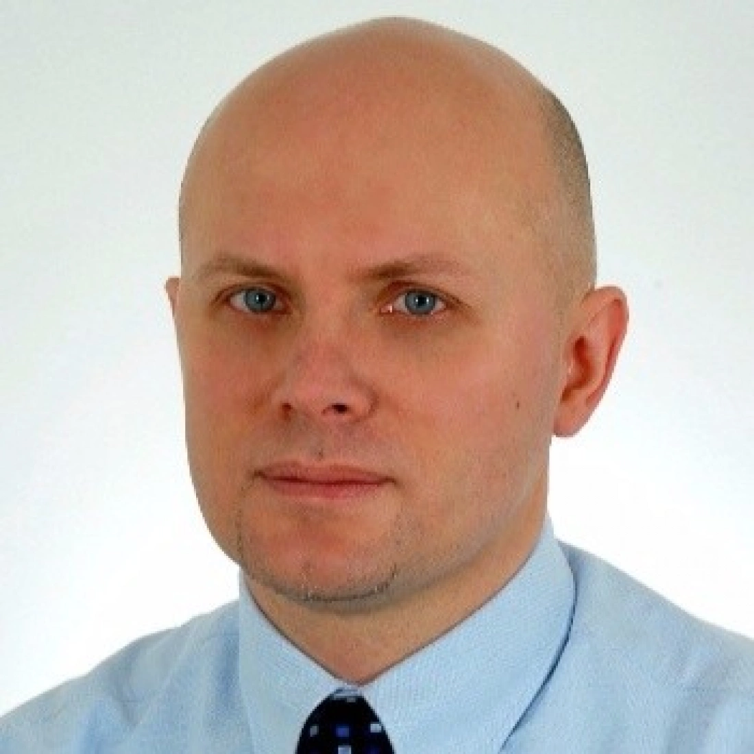 Mężczyzna w jasnej koszuli z krawatem, o łysiejącej głowie i niebieskich oczach, patrzy w kamerę.