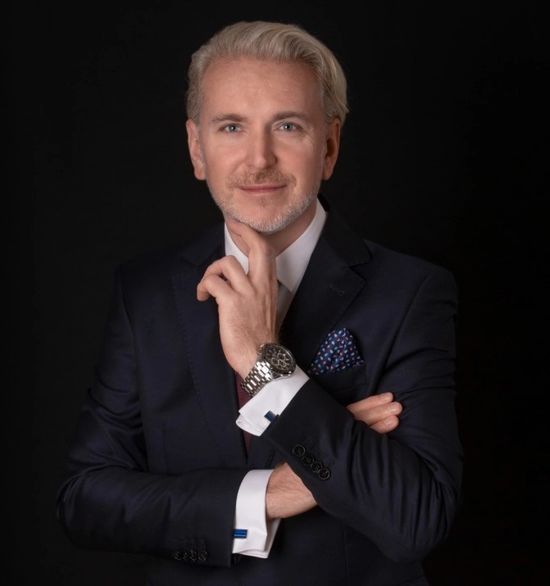 Mężczyzna w eleganckim garniturze, z uśmiechem i ręką przy brodzie, na tle czarnego tła.