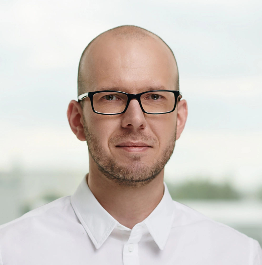 Mężczyzna z okularami i łysiną, ubrany w białą koszulę, pozujący na tle jasnego tła.