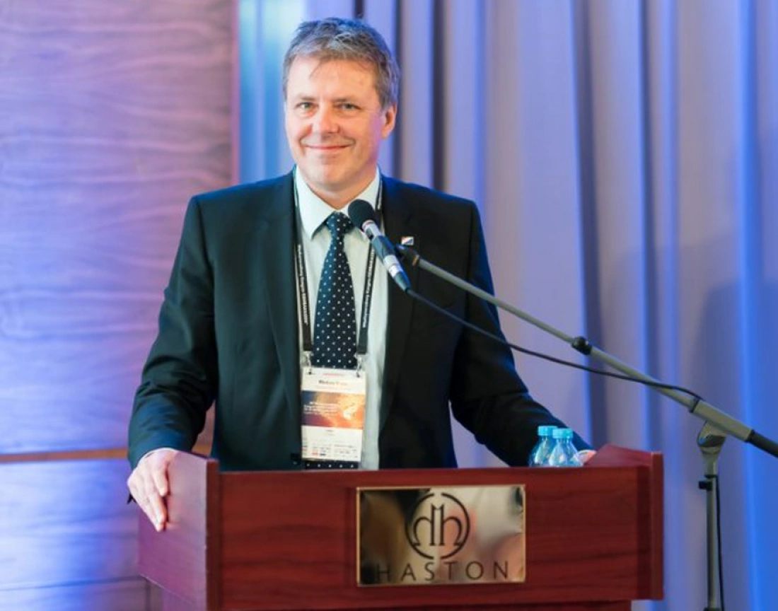 Mężczyzna w garniturze stoi przy mównicy, uśmiechając się do kamery.