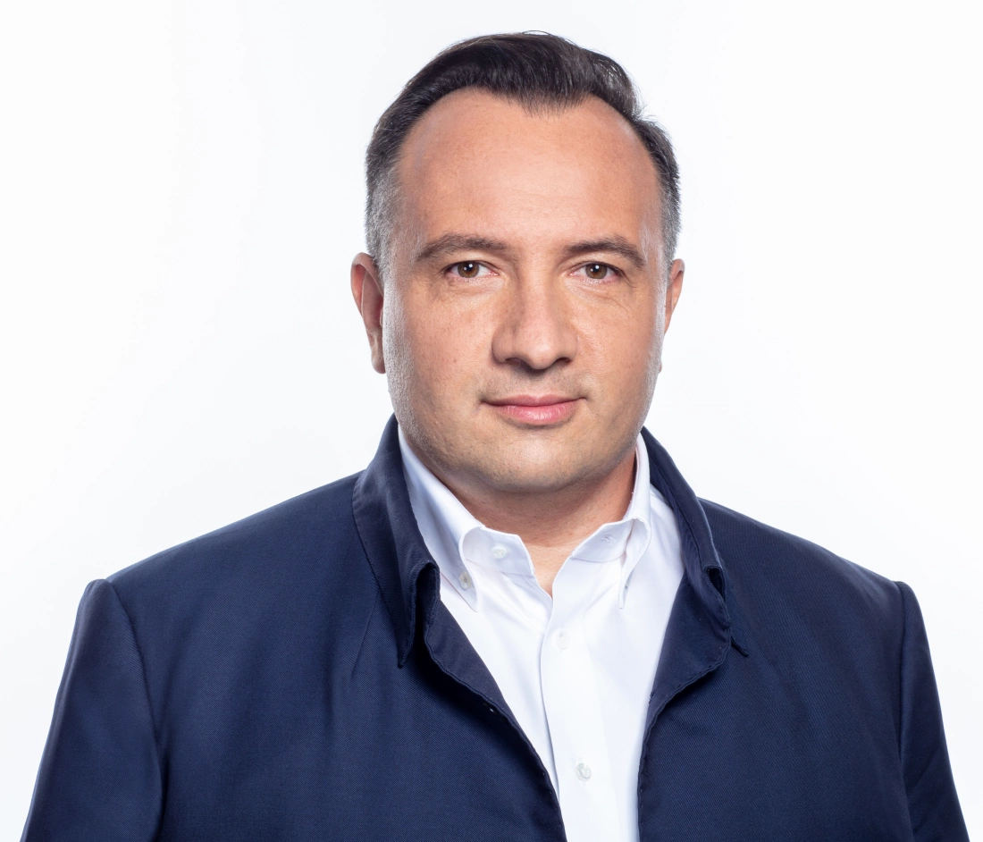 Mężczyzna w jednolitym ubraniu, z poważnym wyrazem twarzy, na jasnym tle.