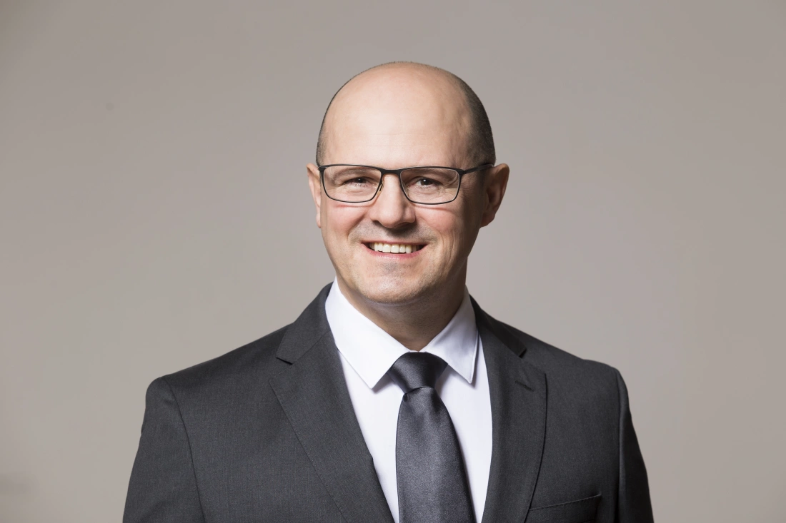 Mężczyzna w eleganckim garniturze z okularami, uśmiechający się na tle szarej ściany.