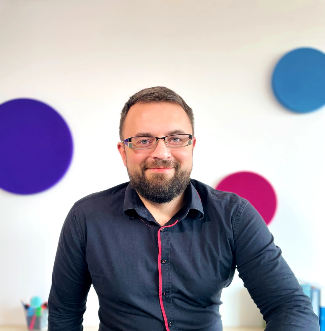 Mężczyzna z brodą w czarnej koszuli stoi przed jasnym tłem z kolorowymi okręgami.