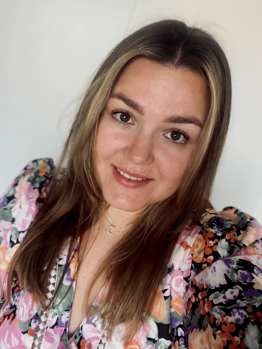 Młoda kobieta uśmiecha się w kolorowej kwiatowej sukience, na tle jasnej ściany.