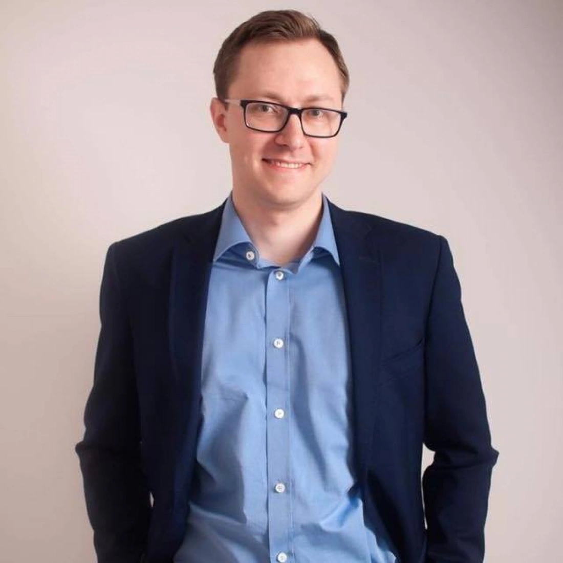 Mężczyzna w okularach, uśmiechający się, ubrany w niebieską koszulę i granatową marynarkę, stoi w neutralnym tle.