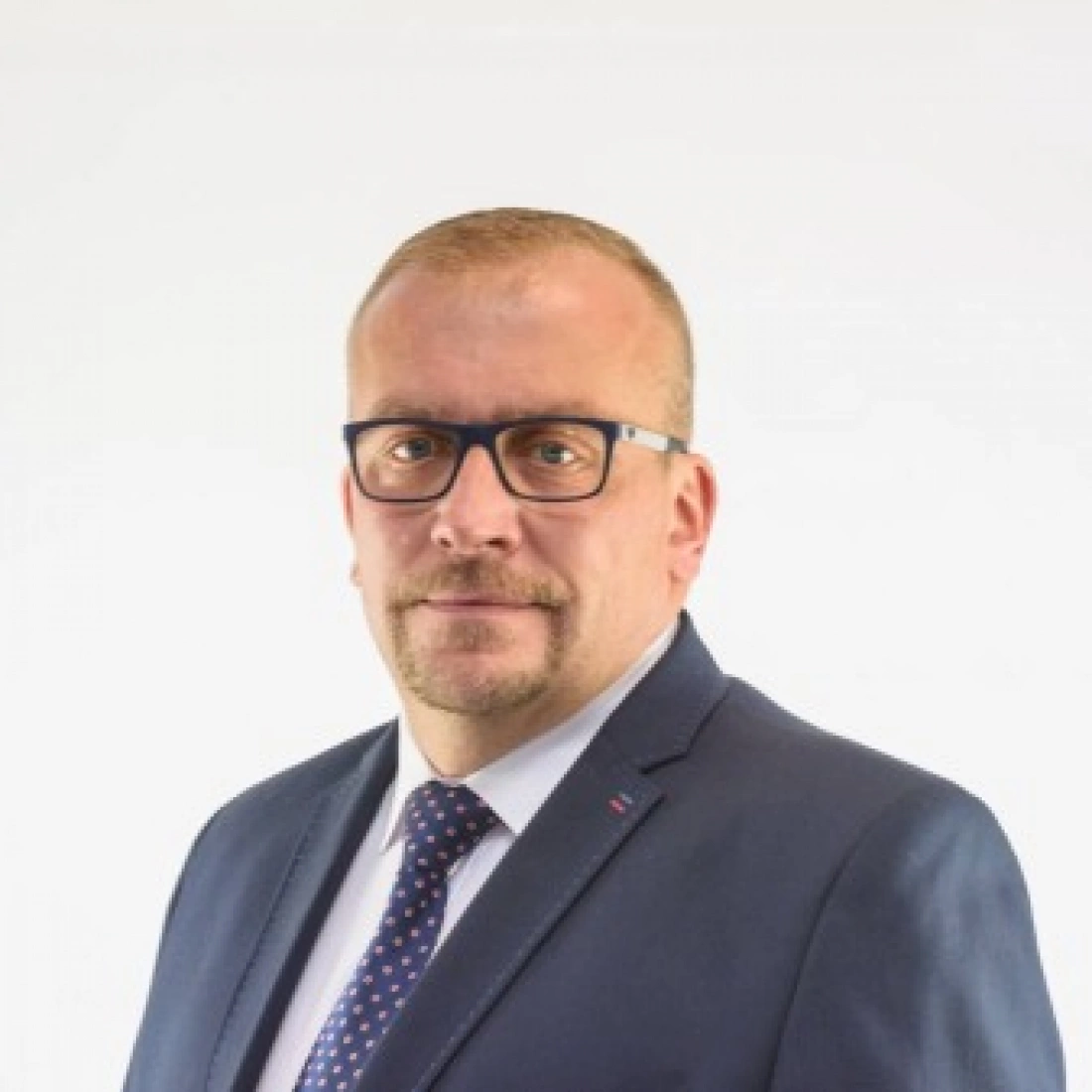 Mężczyzna w garniturze i okularach, z poważnym wyrazem twarzy, na jasnym tle.