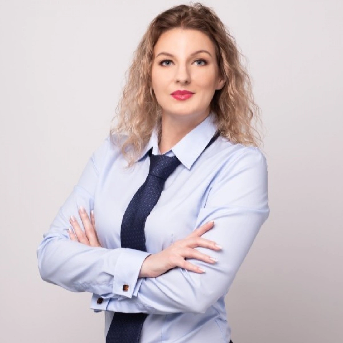 Kobieta w niebieskiej koszuli i krawacie, z założonymi rękami, o falujących włosach.