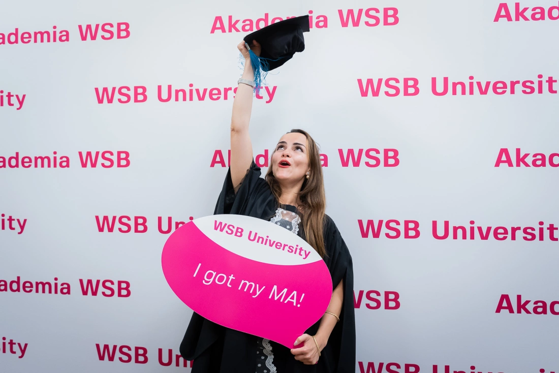 Młoda kobieta w togach akademickich trzyma w górze czapkę oraz transparent z napisem „I got my MA!” przed tłem z logo WSB University.
